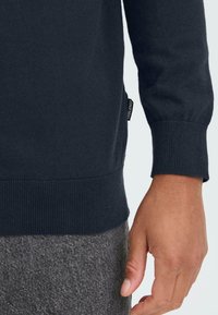 Pull en maille bleu marine avec des poignets et un ourlet côtelés. Il présente une petite étiquette noire sur le côté, associé à un pantalon gris texturé.