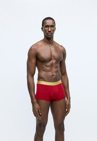 Boxers rouges pour hommes avec une ceinture texturée en or. Tissu lisse, design ajusté et longueur de jambe courte. Le modèle se tient devant un fond neutre.