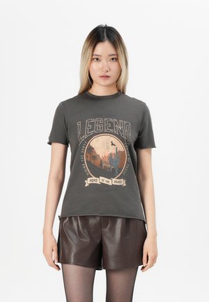 Mujer con cabello largo, vistiendo una camiseta gráfica oscura y pantalones cortos de cuero marrón, de pie frente a un fondo blanco liso.