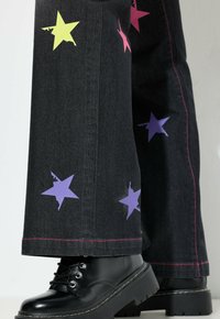 Vaqueros anchos negros con apliques de estrellas coloridas en rosa, morado y amarillo, con costuras rosas y combinados con botas de plataforma negras.