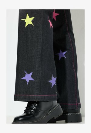 Weite schwarze Jeans mit bunten Stern-Applikationen in Pink, Lila und Gelb, mit pinkem Nähen, kombiniert mit schwarzen Plattformstiefeln.