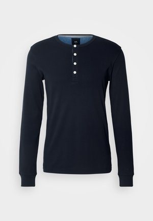 Temno mornarsko modra dolgosleeve majica iz bombaža, z henley ovratnikom in petimi belimi gumbi ter rebrastimi manšetami. Mehka tekstura in prilegajoča zasnova.