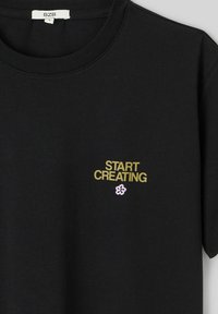 T-shirt noir à col rond, avec l'inscription « START CREATING » en jaune et un petit motif de fleur rose sur le côté gauche de la poitrine.