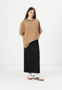Smeđa oversize majica s Adidas logotipom, uparena s crnom maxi suknjom s bočnim trakama i bijelim tenisicama s crnim detaljima.