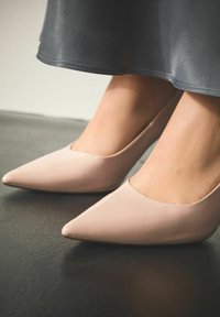 Lichtroze pumps van lakleer met een puntige neus en een gladde afwerking, gecombineerd met een vloeiende, doorschijnende donkergrijze rok, tentoongesteld tegen een donkere vloer.