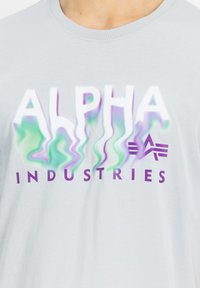 Šedé bavlněné tričko s nápisem "ALPHA" v gradientním efektu zelené a fialové barvy nad "INDUSTRIES" v pevných fialových písmenech. Standardní kulatý výstřih.