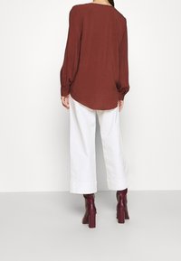 Blouse marron à manches longues avec une coupe décontractée, un ourlet arrondi et des manches bouffantes. Associée à un pantalon large blanc et des bottines à talons bordeaux.