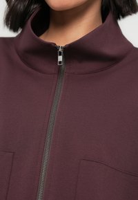 Bordowa kurtka na zamek wykonana z gładkiego materiału, z wysokim kołnierzem, srebrnym zamkiem błyskawicznym i dwoma przednimi kieszeniami dla dodatkowego detalu.