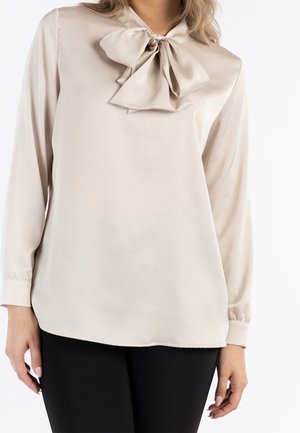 Kobieta ubrana w beżową, satynową bluzkę z długim rękawem i dużą kokardą przy dekolcie oraz czarne spodnie, stoi z ramionami opuszczonymi.