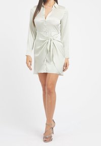 Nainen, jolla on päällään vaaleanvihreä satiinimekko, jossa on pitkät hihat, etunapit ja solmittava vyö, yhdistettynä hopeisiin korkokenkiin.