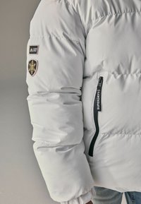 Doudoune blanche avec des poignets côtelés, zip latéral et écussons présentant un flocon de neige et le logo de la marque. Texture du matériau lisse et brillante.