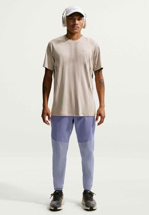 Mężczyzna w beżowej czapce, bezprzewodowych słuchawkach, beżowej sportowej koszulce, dwukolorowych fioletowych spodniach i czarnych butach do biegania, stojący na białym tle.