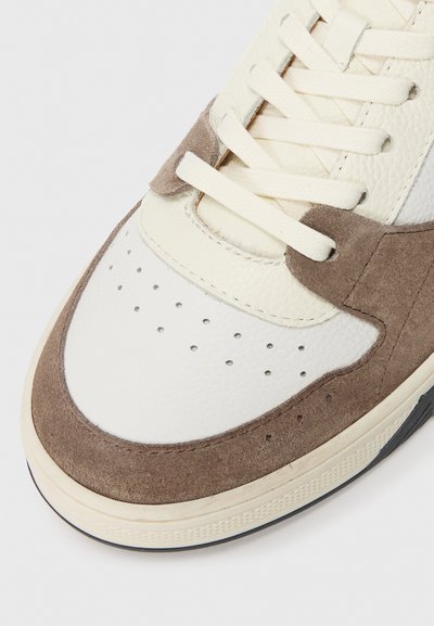 Närbild av en beige och vit sneaker med perforerat läder, mockaöverdrag och gräddfärgade snören på en ljus bakgrund.