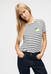 Νεαρή γυναίκα με μακριά ξανθά μαλλιά, φορώντας μια λευκή και μαύρη ριγέ μπλούζα με κίτρινο μπαλώμα λεμονιού και σκούρο μπλε τζιν, με τα χέρια στις τσέπες.