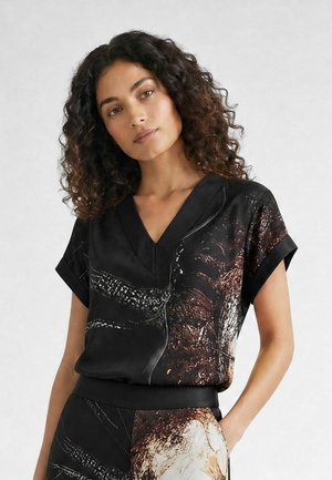 Femme aux cheveux bouclés portant un haut noir à manches courtes en V avec des motifs abstraits marron et beige, debout avec une main dans la poche.