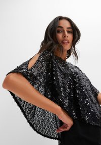 Top nero con paillettes, a maniche corte, con spalle scoperte e vestibilità ampia. Il tessuto è lucido e testurizzato, riflettendo la luce.