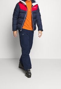 Marineblaue Pufferjacke mit roten und weißen Akzenten, orangefarbenes Oberteil, marineblaue Hose und schwarze Stiefel. Strukturiertes Material mit Reißverschlusstaschen an der Hose.
