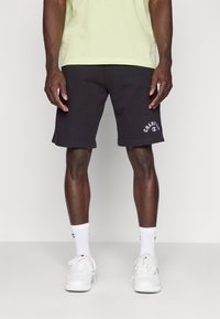 Svarta bomullsshorts med ribbad midja och vitt logodetalj på vänster ben, som kombineras med vita sneakers och strumpor.
