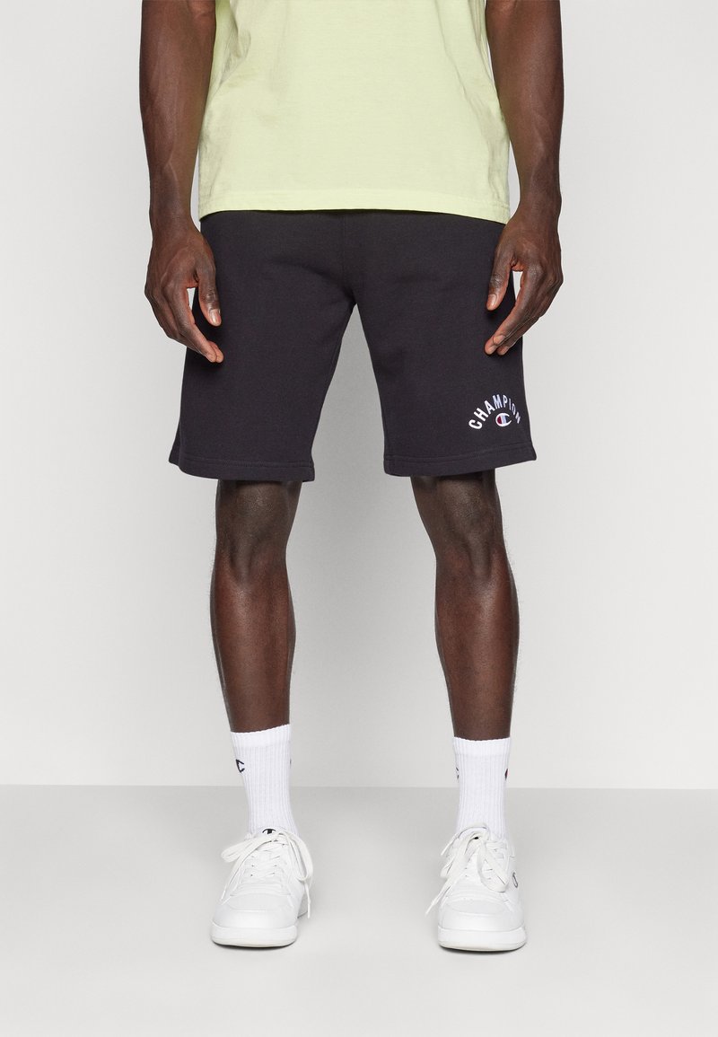 Svarta bomullsshorts med ribbad midja och vitt logodetalj på vänster ben, som kombineras med vita sneakers och strumpor.