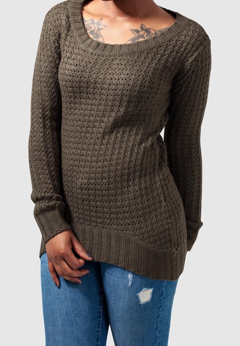 Femme portant un pull en maille marron texturé avec un décolleté en forme de creux et un jean bleu déchiré, montrant des tatouages floraux sur la partie supérieure de la poitrine.