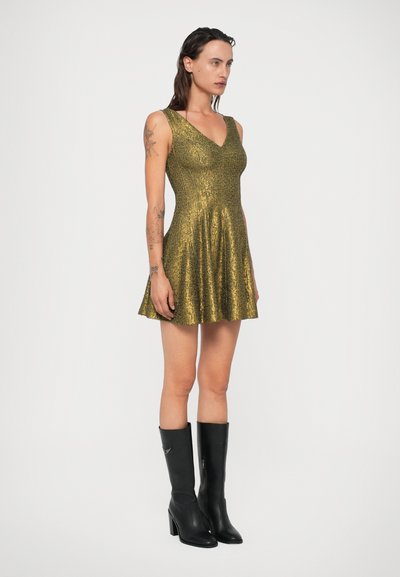 Auksinių blizgučių suknelė su platėjančia sijonu, V formos iškirpte ir plonais petnešomis; derinama su juodais aukštakulniais batais iki kelių. Lygus paviršius.