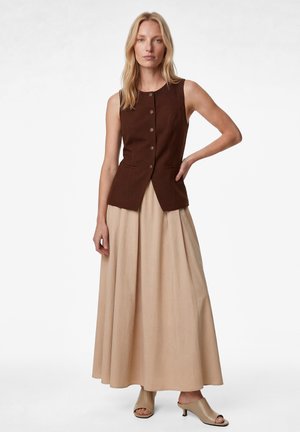 Frau mit blondem Haar, die eine ärmellose braune Weste mit Knöpfen, einen langen beigen Rock und beige offene Absatz-Mules trägt, steht mit einer Hand in der Hüfte.