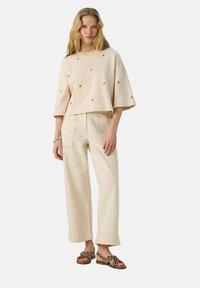 Haut à manches courtes en crème avec broderie motif goutte orange, associé à un pantalon large beige et des sandales imprimé léopard.