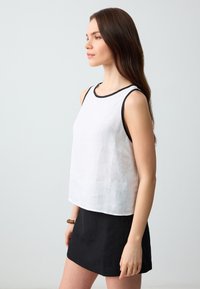 Haut sans manches en lin blanc avec un col noir, associé à une mini jupe noire. Le tissu semble léger et texturé. Design simple et décontracté.