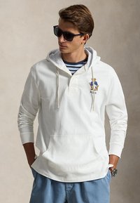 Jeune homme portant des lunettes de soleil, un sweat à capuche blanc avec un logo ours, une chemise rayée en dessous, un pantalon bleu et une montre-bracelet, debout les mains dans les poches.