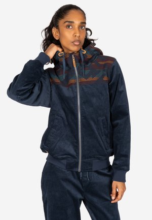 Person wearing a navy blue zip-up hoodie with patterned upper chest and hood, paired with matching navy blue pants.