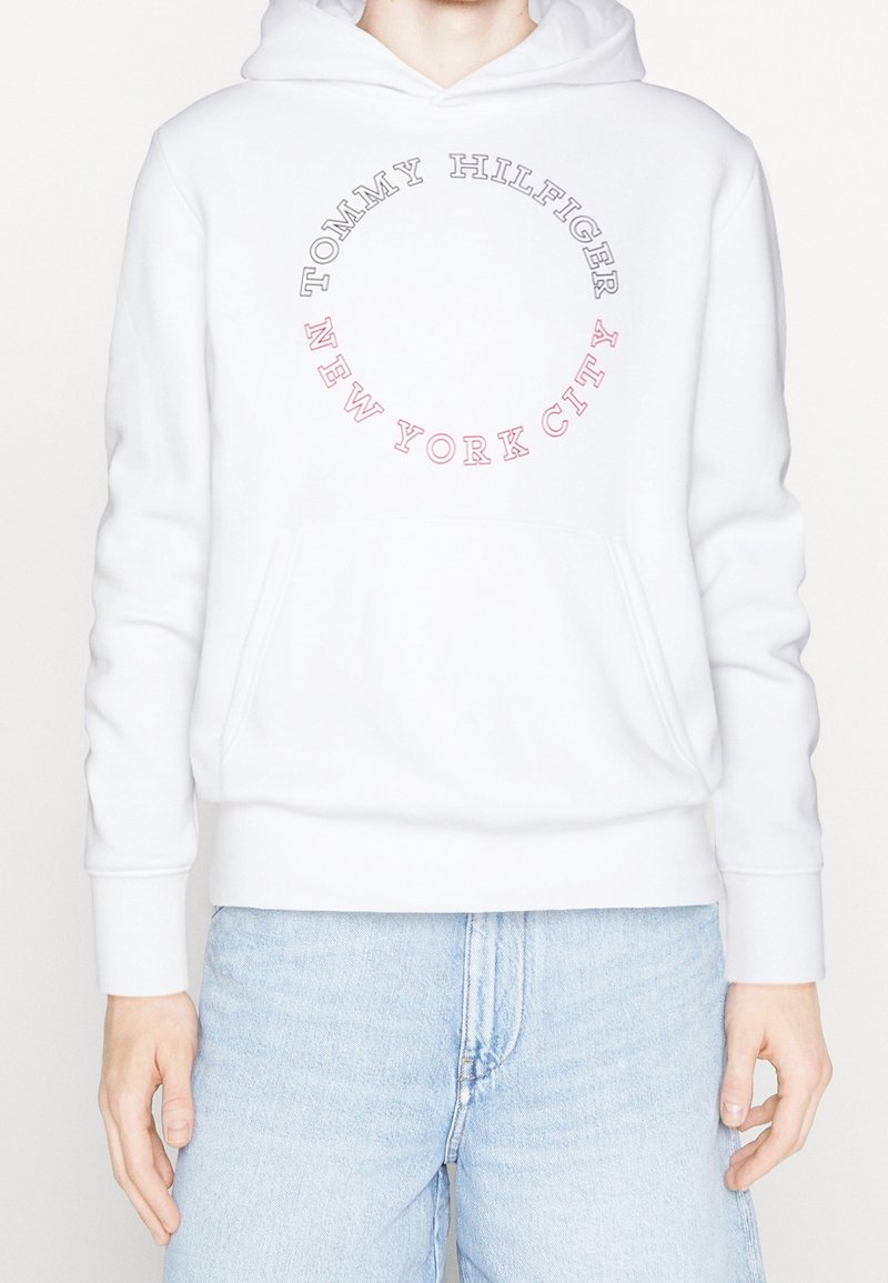 Sweat à capuche blanc avec texte circulaire « Tommy Hilfiger New York City » en noir et rouge, porté avec un jean en denim bleu clair.