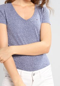 Gråblå, kortärmad t-shirt med V-hals och mjuk textur; har en liten logotyp på bröstet och är kombinerad med vita byxor.