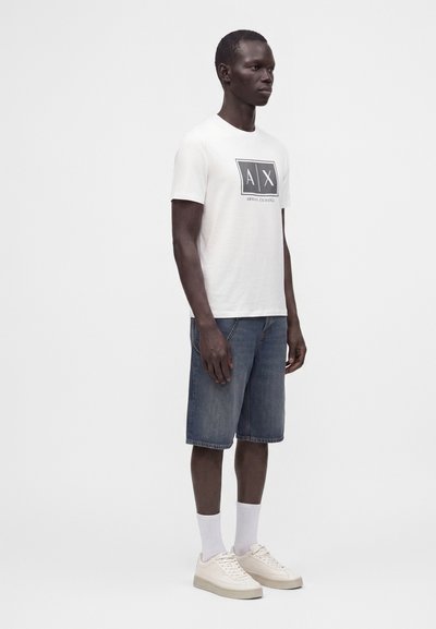 Мъж, облечен с бяла тениска с лого на Armani Exchange, сини дънкови къси панталони, бели чорапи и светлобиежови маратонки на неутрален фон.