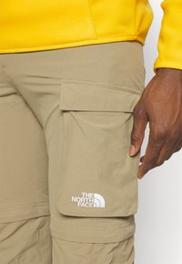 Beige cargobyxor med sidfickor, tillverkade av lätt material. Har en vit logotyp: "THE NORTH FACE" nära fickan.