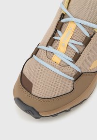 Αθλητικό παπούτσι σε μπεζ χρώμα με πλέγμα στην επάνω επιφάνεια, ανοιχτά μπλε και κίτρινα κορδόνια και καφέ λεπτομέρειες. Διαθέτει ενισχυμένο μπροστινό μέρος και υφή στη σόλα.