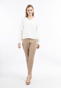 Witte, gebreide V-hals trui gecombineerd met beige tapered broeken en nude hakken. Model staat tegen een witte achtergrond.