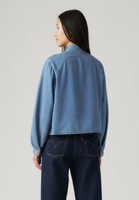 Light blue denim jacket with long, puffed sleeves and a cropped design. Features buttoned cuffs and a row of stitching along the shoulders.