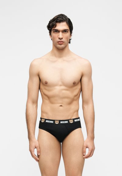 Νέος άνδρας με γυμνασμένο σώμα που φοράει μαύρα εσώρουχα με αρκουδάκι και ελαστική μέση με το λογότυπο "Moschino", στέκεται μπροστά σε απλό φωτεινό φόντο.