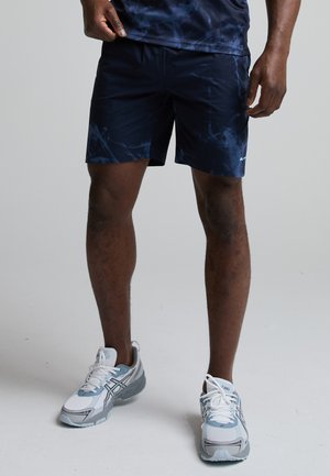 Mand iført marineblå mønstrede sportsshorts og hvide og grå løbesko, stående på en ensfarvet lys baggrund.
