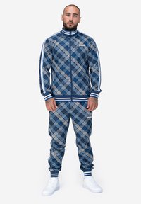 Tamsiai mėlynas treningas su baltu languotu raštu, turintis užtraukiamą švarką su aukštu apykakle ir atitinkančias siauras kelnes, abi su juostuotais rankogaliais.