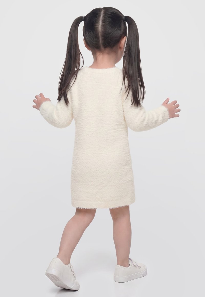 Jeune fille aux couettes foncées portant une robe crème duveteuse et des baskets blanches, debout et de dos sur un fond clair uni.