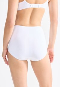 Vrouw in witte hooggewaiste slip en een bijpassende witte beha, van achteren te zien tegen een effen witte achtergrond.