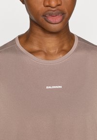 Καφέ διαπνέον μπλουζάκι με στρογγυλή λαιμόκοψη και λευκό λογότυπο "SALOMON" στο στήθος. Το ύφασμα έχει υφή με οπτική αίσθηση.