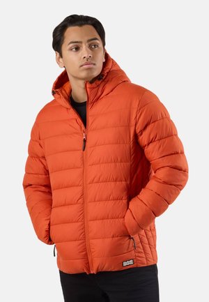 Giovane uomo con giacca trapuntata arancione brillante con cappuccio, mani nelle tasche, che guarda leggermente di lato su uno sfondo chiaro e uniforme.