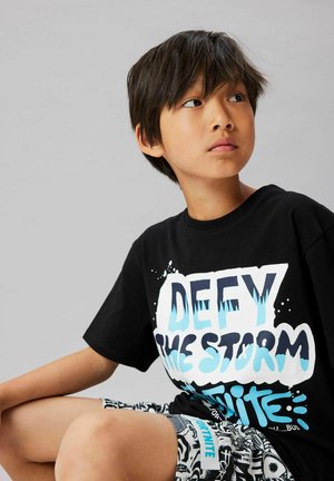 Giovane seduto, indossa una maglietta nera con la scritta "Defy the Storm Fortnite" e pantaloncini a fantasia, guarda di lato su uno sfondo semplice.