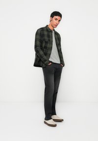 Ung mann står med hendene i lommene iført en grønn og svart rutete jakke, grå skjorte, svarte jeans og beige joggesko mot en hvit bakgrunn.
