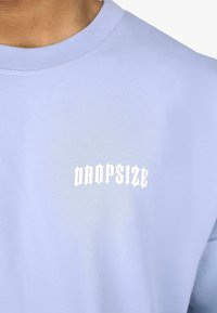 Lys blå bomullssweatshirt med rund hals og hvit "DROPSIZE" tekst i en fet, stilisert skrift på venstre brystområde.