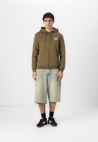Olivnozeleni zip-up hoodie s vezicami, sprednjimi žepi in detajlom logotipa. V kombinaciji z svetlo modrimi kavbojkami in črnimi supergami.