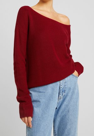 Kvinde iført en burgunderfarvet off-shoulder striktrøje og lyseblå højtsiddende jeans med den ene hånd i lommen foran en ensfarvet baggrund.