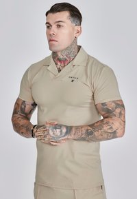 Beżowa koszulka polo z krótkim rękawem, z dekoltem w serek, logo na piersi i dopasowanym krojem. Wykonana z gładkiego, elastycznego materiału.